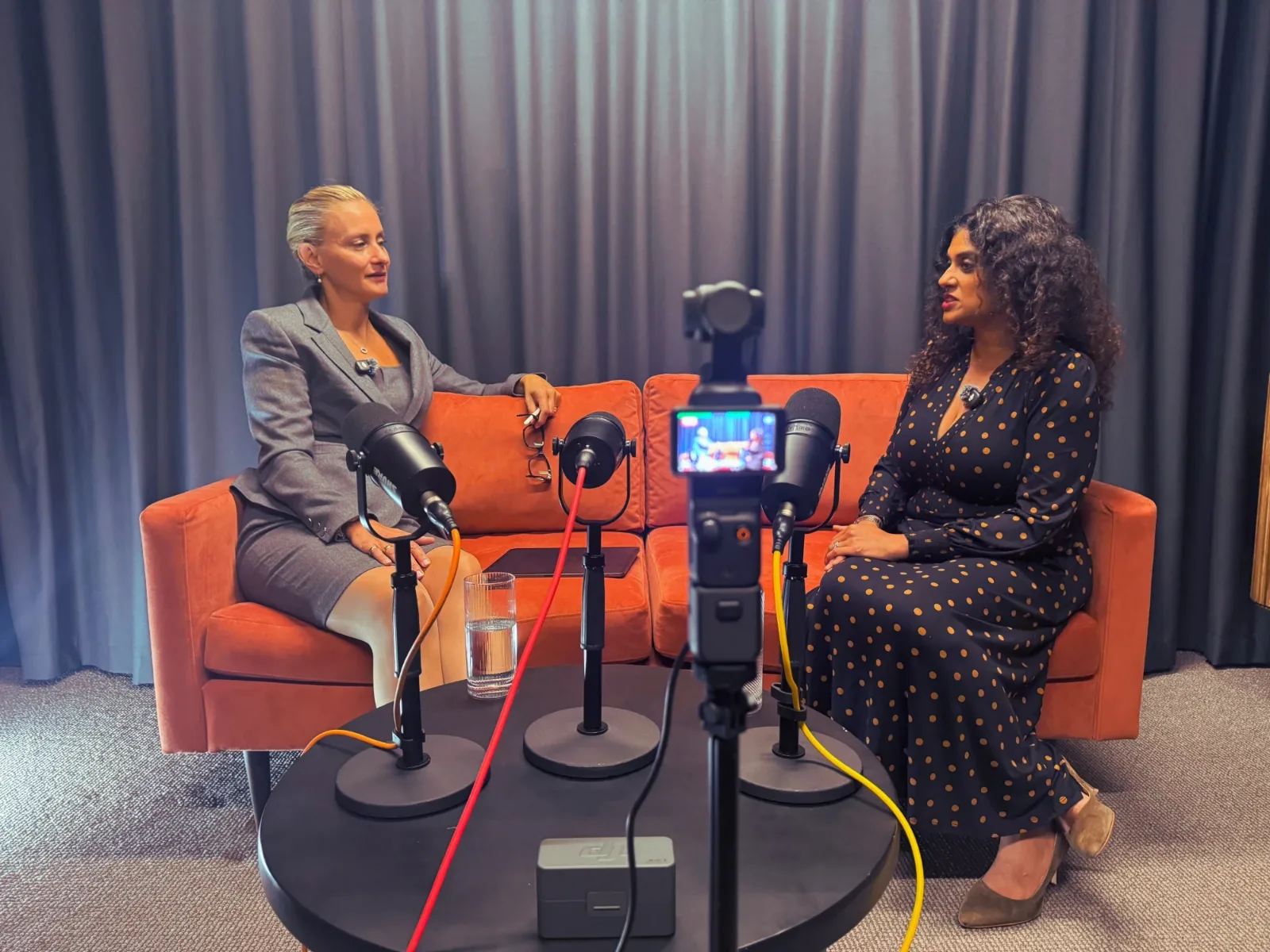 Two women in conversation on an orange sofa at Great Portland Street Studio, with three podcast microphones on a round table and a camera filming in the foreground