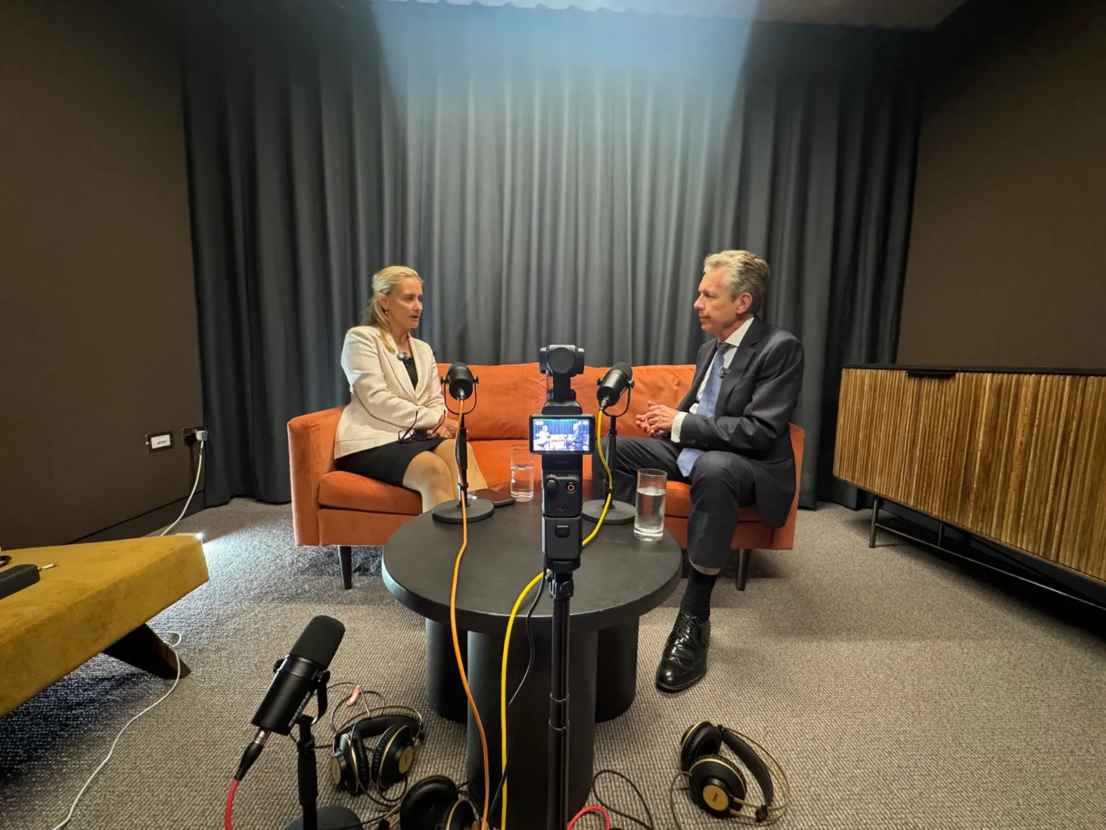Two people in conversation on an orange sofa at Hale House, filmed on a gimbal camera with podcast microphones and headphones set up around them