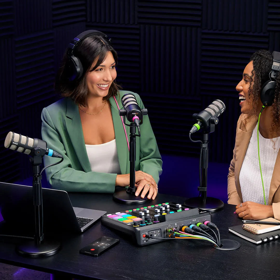 Two podcast hosts in headphones laughing across a desk with RØDE microphones and a RØDECaster Pro II mixer